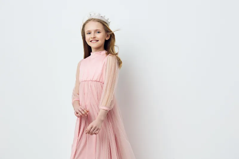 Smiling girl in a pink dress and crown, exuding joy and innocence against a soft white background, perfect for children s fashion or celebration concepts