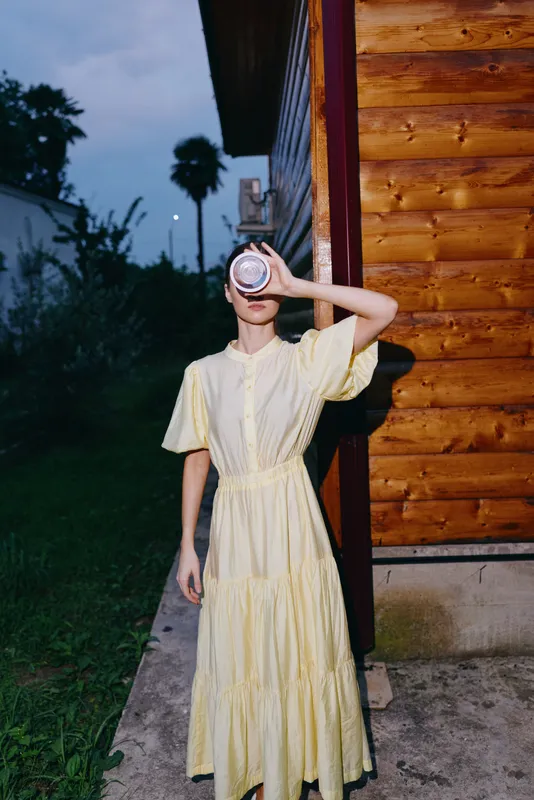 Person wearing a light yellow dress stands beside a wooden wall at dusk, covering eyes with a round object, creating a moody outdoor portrait.