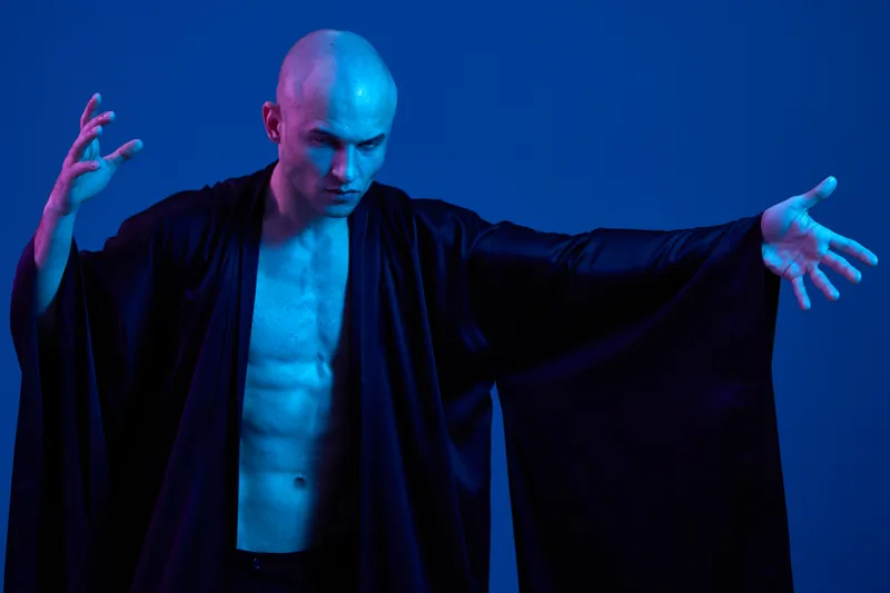 Intense man in a black robe exudes power and confidence, showcasing a strong pose against a vivid blue background. Dramatic lighting enhances the atmosphere.