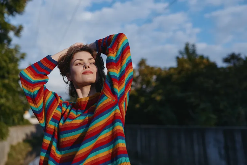 Woman portrait in colorful rainbow sweater outdoors, relaxed and smiling with hands behind head, casual fashion lifestyle, natural light, autumn trees and clear sky background.