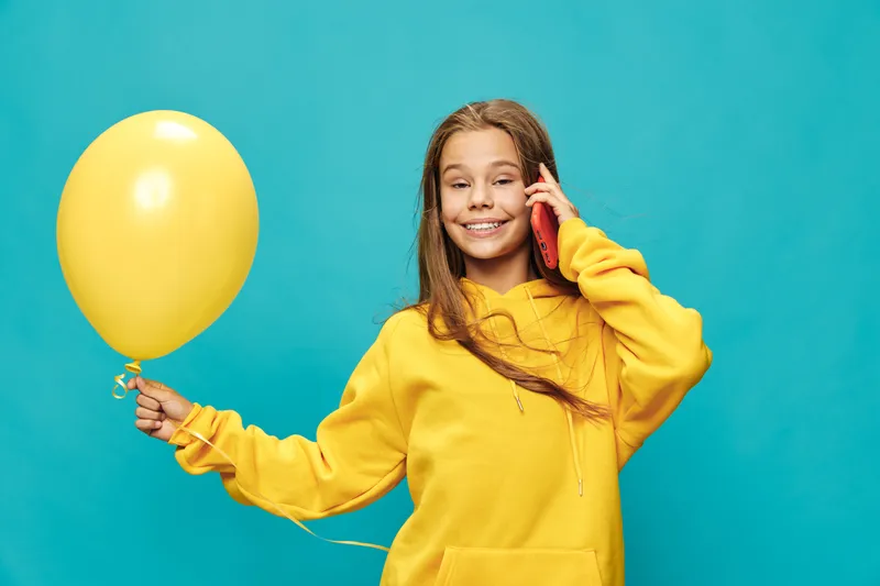Girl in yellow hoodie holds yellow balloon and talks on mobile phone, smiling against blue background. Child with long hair in casual clothes for childhood, communication and fun.