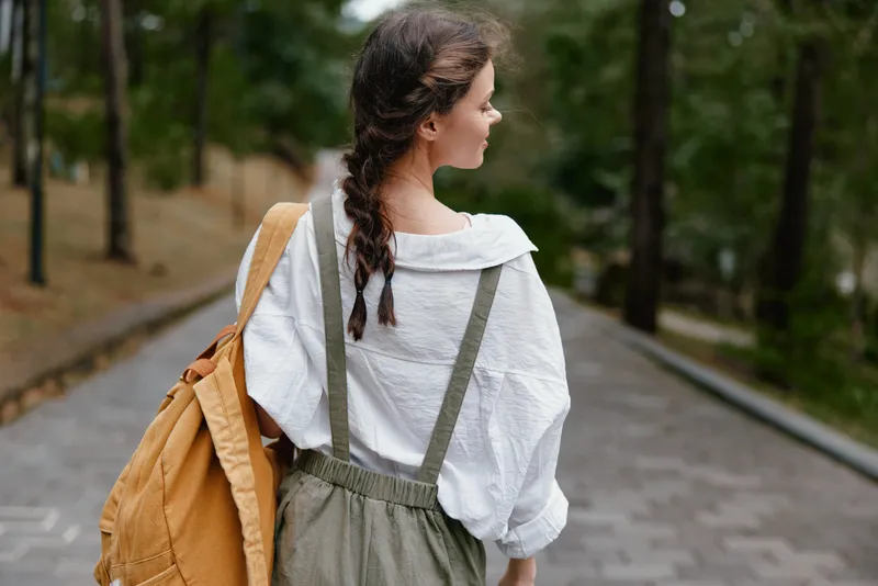 Young woman in casual outfit with backpack walking in a serene park, showcasing a relaxed vibe, ideal for themes of travel, nature, and adventure