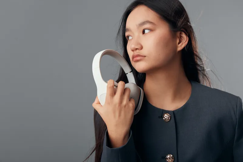 Young woman holding headphones with one hand looking sideways on gray background, fashion portrait with natural makeup and long dark hair in modern jacket in studio light.