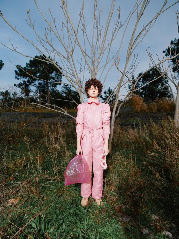 Pink jumpsuit worn by a woman in an outdoor field with a bare tree, carrying a pink bag, moody light and rustic natural backdrop during golden hour.