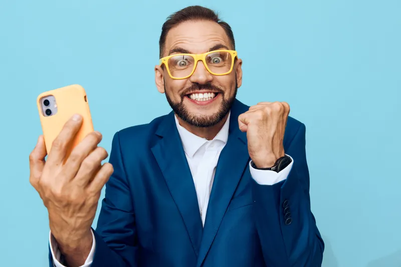 A professional man in a blue suit takes a selfie with yellow glasses, standing against an isolated colored background, displaying confident and joyful emotions.
