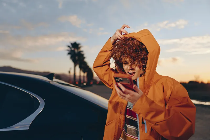 Woman lifestyle portrait, curly hair under hooded orange jacket, smiling, holding phone near car at sunset outdoors, palm trees, warm evening light, casual travel, communication, city road