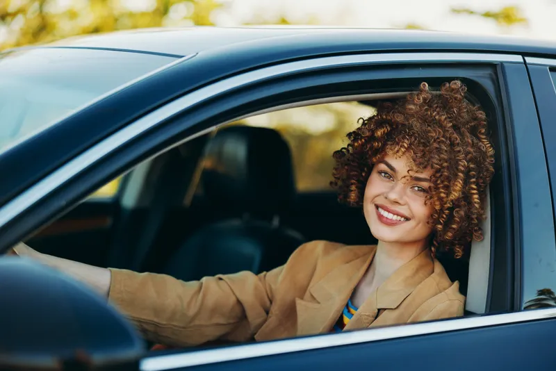 Smiling woman with curly hair driving a car, wearing a tan blazer. Warm, retro tones create a cozy mood, highlighting modern lifestyle, travel, and daily confidence behind the wheel.