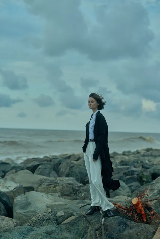 Woman stands on jagged rocks by the sea, wearing a long coat and pale trousers, wind blowing her hair, moody coastal scene, dramatic fashion moment captured outdoors at dusk