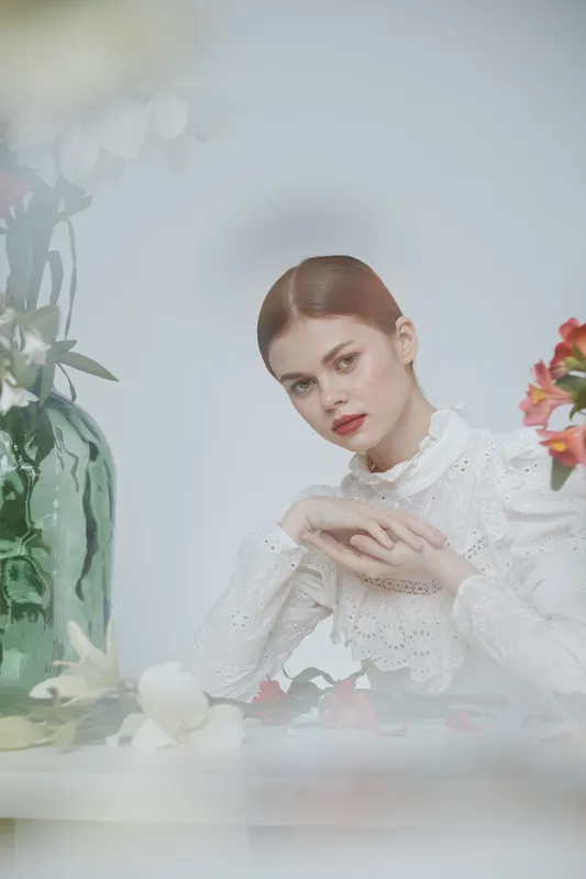 Elegant young woman in white lace blouse, surrounded by flowers, showcasing a serene expression, bright colors, and soft background, perfect for beauty and fashion themes