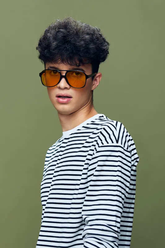 Young man with curly hair wearing orange sunglasses and a striped shirt posing against an olive green background, showcasing contemporary fashion style.