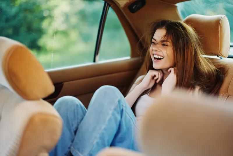 Happy young woman laughing in a car, wearing casual jeans and a white top, enjoying a sunny day, creating a joyful and carefree atmosphere.