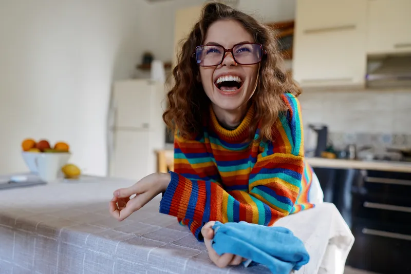 Joyful woman in a colorful striped sweater laughs at a kitchen table, a candid home moment that conveys happiness, warmth, and casual daily life. Natural light, relaxed atmosphere, candid.