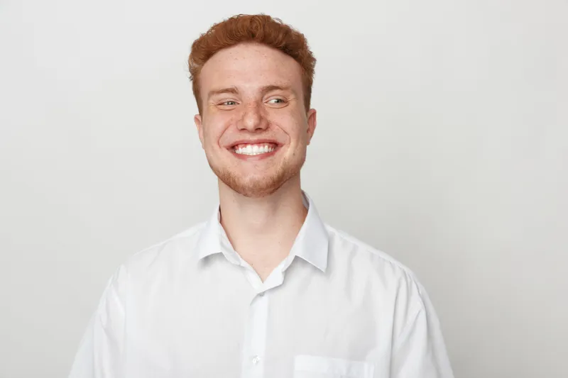 Smiling young man with red hair and beard wearing white shirt looks to side on light background. Concept of happiness, confidence, casual style, and positive emotions in portrait photography.