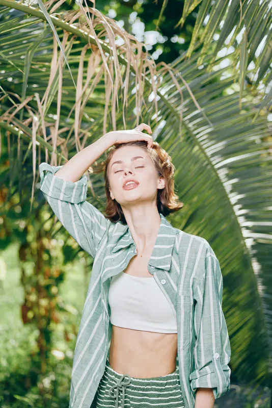 Sunlit portrait of a young woman outdoors, wearing a striped shirt over a white crop top, posing confidently amid tropical foliage.