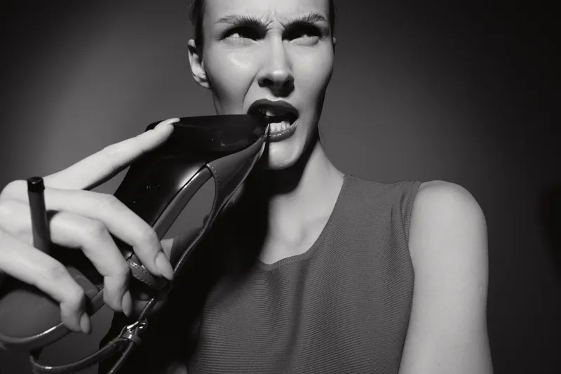 Young white woman with pale skin bites black high heel shoe with frustrated expression in studio.