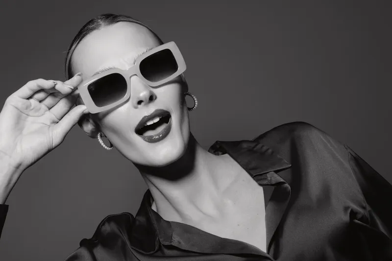 Stylish white woman with light skin wearing sunglasses and dark satin shirt poses confidently against dark background in studio portrait.