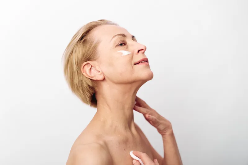 Mature woman with short hair applies white cream on face and neck, showing skincare routine and self-care concepts on bright white background. Focus on healthy skin and beauty.