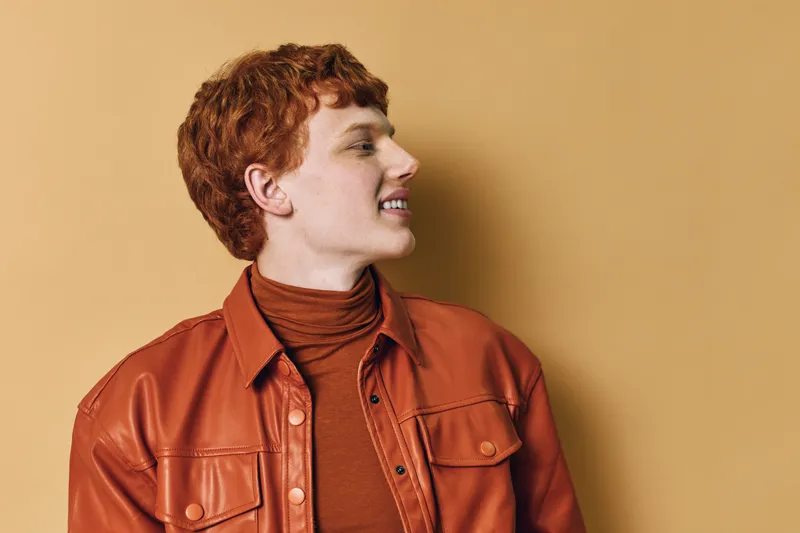 Young man with red hair wearing a brown leather jacket and turtleneck looking sideways smiling against a beige background in studio portrait.