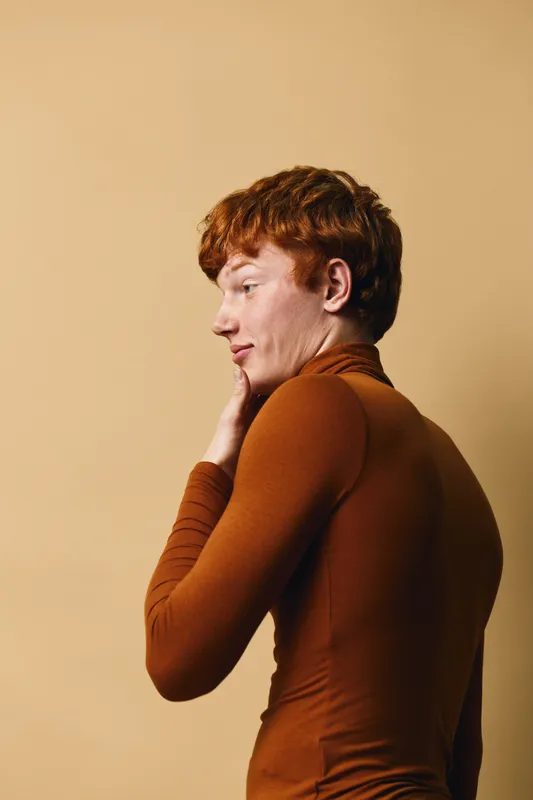Young man with red hair wearing brown turtleneck standing sideways on beige background with hand near face, thoughtful expression and casual style portrait.