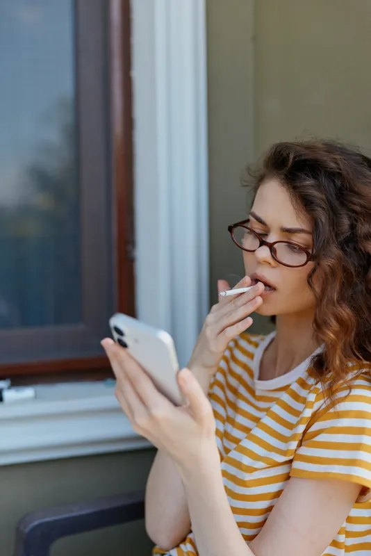 A woman in a striped t shirt and glasses sits on a balcony, scrolling on a smartphone and tasting a snack. A casual, everyday home lifestyle moment reflecting personal time.