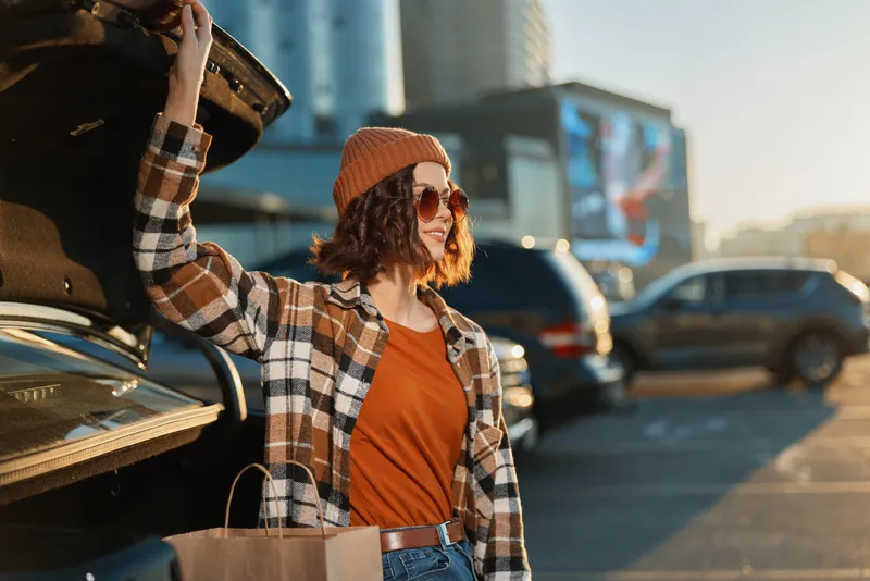 Woman by car trunk with shopping bags at sunset in urban parking, beanie and sunglasses, candid lifestyle portrait with authenticity, golden hour glow and mindful living vibe.