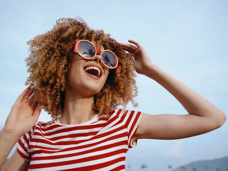 Joyful woman with curly hair wears a red and white striped shirt and round sunglasses, enjoying sunshine and a carefree moment outdoors in bright daylight.