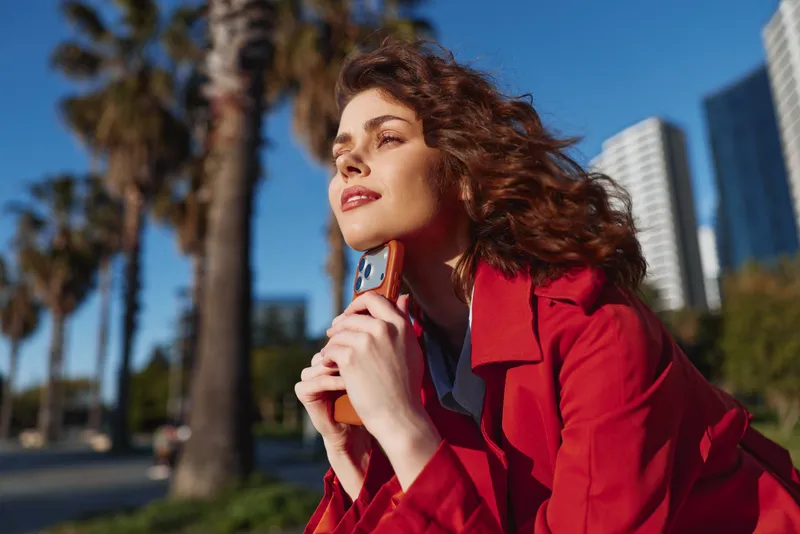 A confident woman in a red coat outdoors poses with a vintage cellphone, gazing toward the distance. Bright sunlight, palm trees, and tall buildings frame an elegant urban portrait.