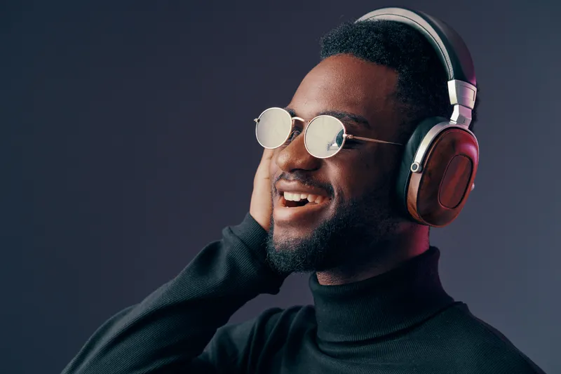 Young Black man in stylish headphones enjoying music in a dark studio, showcasing joy and relaxation Stylish attire, vibrant emotions