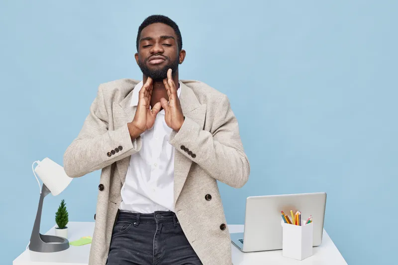 Confident young man relaxing at workspace, stretching neck, eyes closed, wearing beige blazer and white shirt, modern office setting with laptop, desk lamp, and stationery, calm mood. People lifestyle