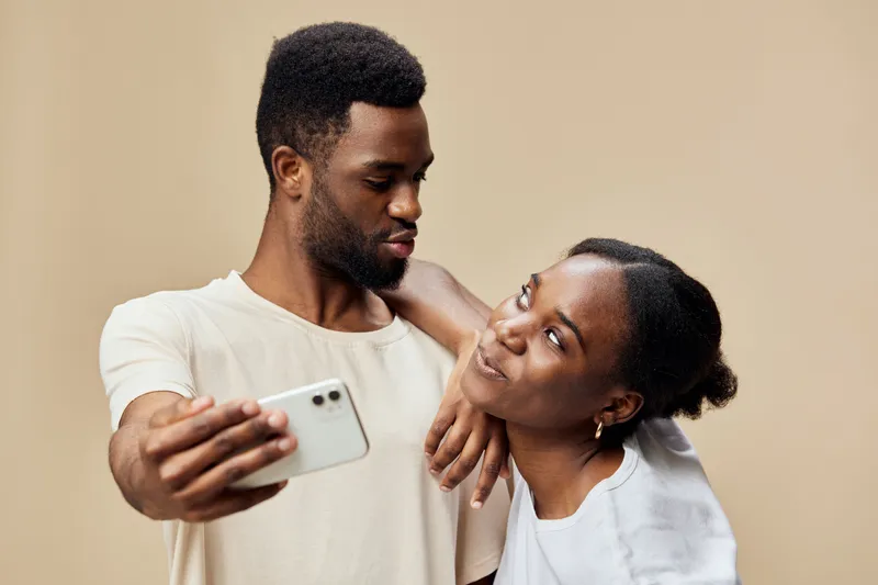 Young couple taking a selfie together, smiling and sharing a joyful moment against a neutral background Their casual outfits reflect a relaxed lifestyle, highlighting love and connection