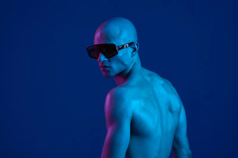 Athletic man with stylish sunglasses striking a pose against a vibrant blue backdrop, showcasing a unique fashion concept and modern aesthetics.