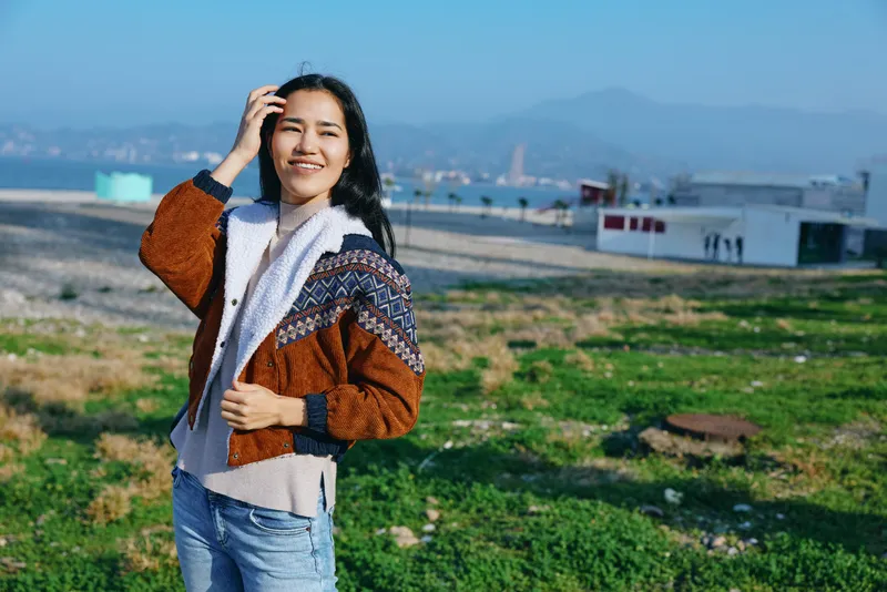 Bright outdoor portrait of a smiling woman in a knitted jacket standing in a grassy field with distant hills under a clear blue sky, showcasing natural style and relaxed travel vibe