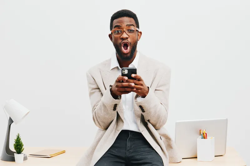 Excited young man expressing surprise while using smartphone in modern home office, wearing casual beige jacket, sitting at desk with laptop, plant, and stationery, white background, lifestyle concept