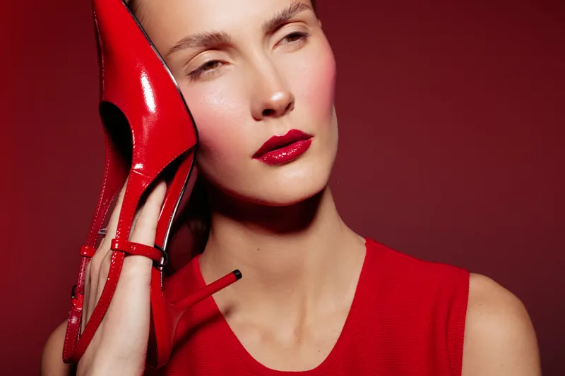 Close-up of a woman with red lipstick holding a red high heel shoe near her face against a red background. Concept of fashion, beauty, style, and elegance in studio lighting.