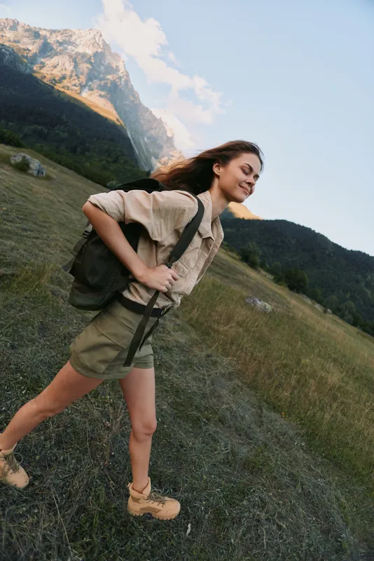 Cheerful woman outdoors, hiking along a grassy slope with a backpack, mountain backdrop, clear sky, sunlit moment, relaxed posture, natural colors, fresh air