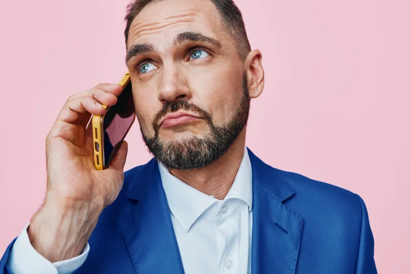 A professional man on a pink studio backdrop speaks on a bold phone, conveying thoughtful emotion. The isolated colored background highlights a confident business vibe and sharp posture.
