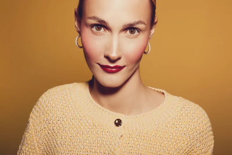 Close-up portrait of a smiling woman with clear skin wearing a knitted sweater and hoop earrings on a warm yellow background. Concept of beauty, fashion, and confidence.