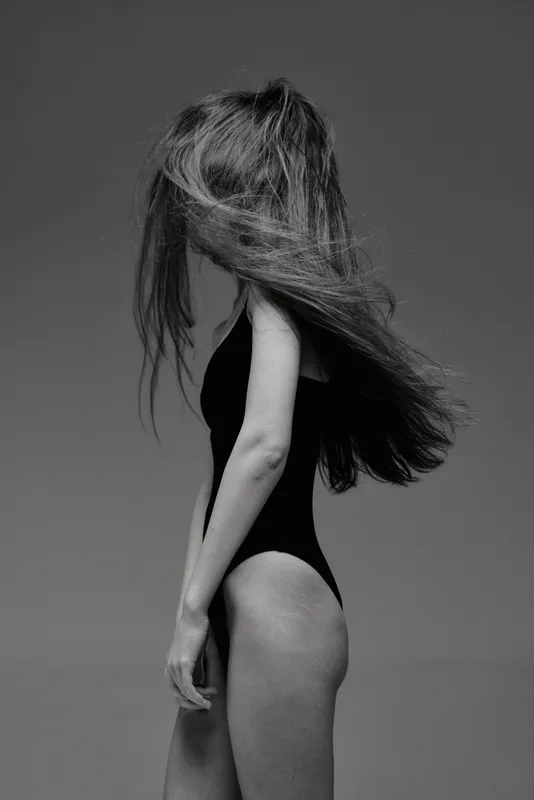Black and white artistic portrait of a woman with long hair in a black bodysuit posing in studio.
