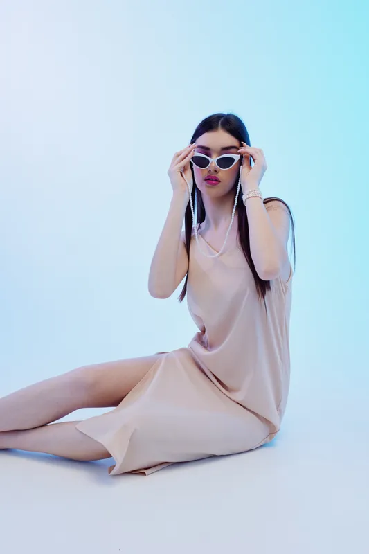 Young woman in a beige dress adjusts white sunglasses while sitting on the floor. Studio shot with soft lighting and a light blue background. Concept of woman beauty fashion.