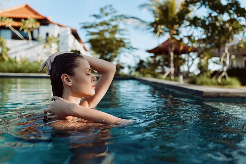 Relaxed woman in a sunlit pool, eyes closed, gentle breeze, tropical resort backdrop, casual pose, clear water, warm colors, carefree mood, lifestyle moment