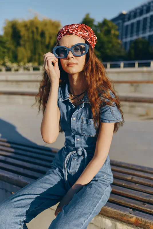 Young woman wearing denim jumpsuit and sunglasses sitting outdoors on a sunny day with urban background and casual style