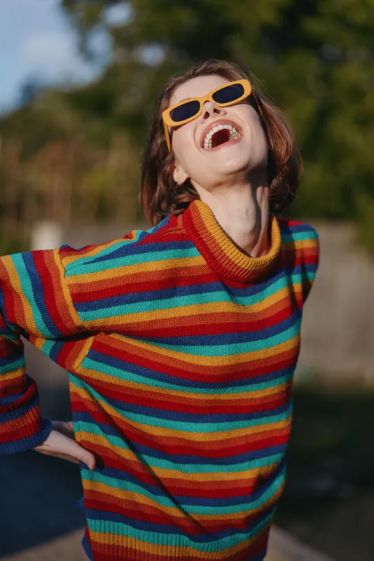 woman smiling striped sweater colorful sunglasses outdoors young adult joyful short hair casual outfit. Young woman in her 20s wearing a rainbow knit, leaning back and laughing in park sunlight for