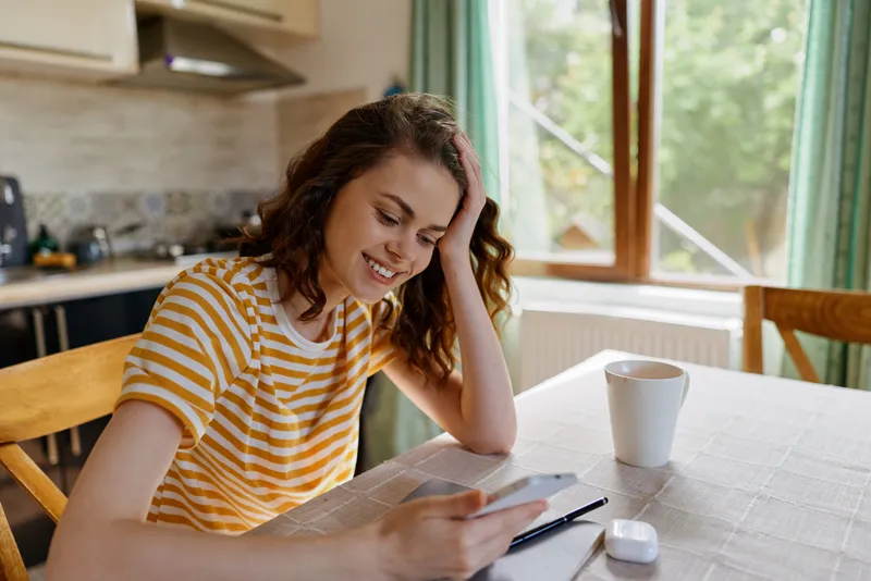 A smiling woman in a striped yellow shirt sits at a table in a sunny kitchen, using a smartphone. A cozy, casual home moment focused on everyday technology and domestic life.