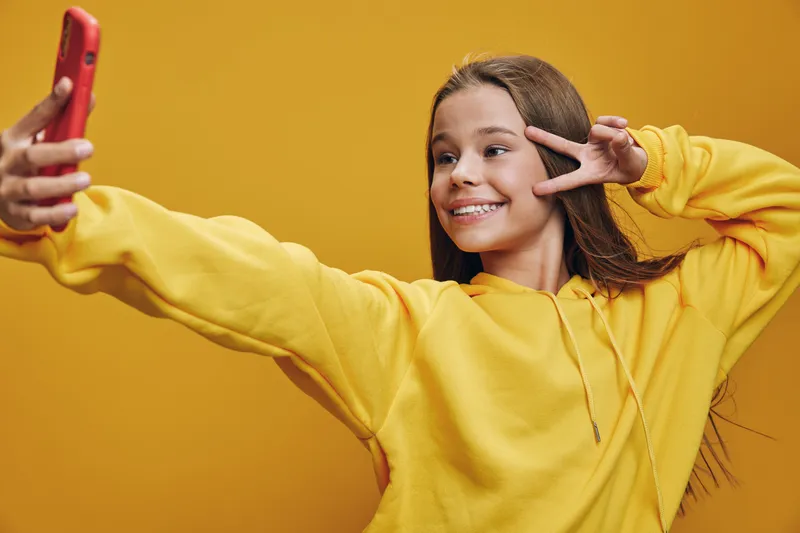 Girl in yellow hoodie takes selfie with smartphone smiling and showing peace sign on yellow background. Youth, happiness, casual and modern digital lifestyle concept.