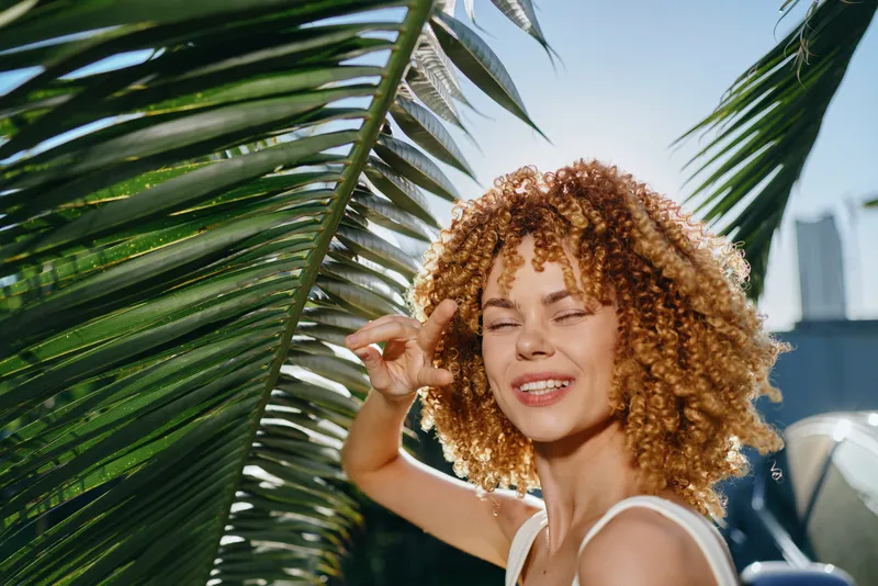 Smiling woman with curly hair enjoys sunny tropical outdoor setting surrounded by green palm leaves and clear blue sky during daytime.