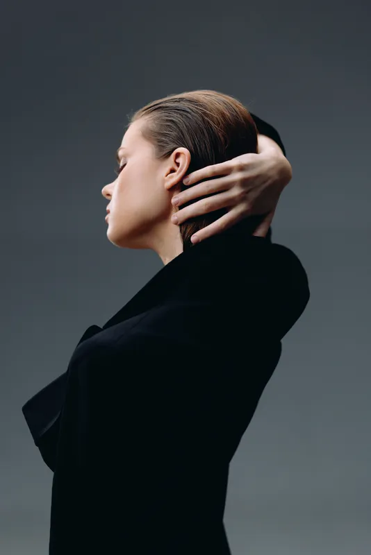 Profile of a thoughtful woman in a black outfit with hand on neck against neutral background