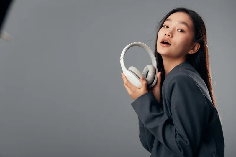 Young woman holding headphones with one hand on gray background. Casual style female model with long hair posing for audio technology or music concept in studio lighting.