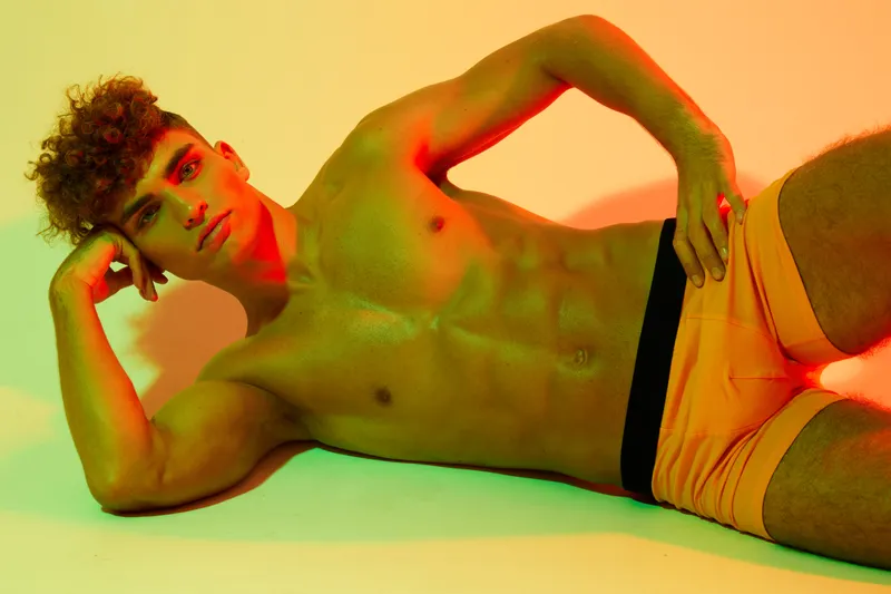 Young man with muscular body lying on side in colorful studio lighting, wearing orange shorts and posing with hand on head for fitness or fashion concept.