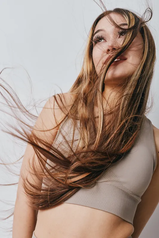 Young woman with long hair flowing, smiling and looking up wearing sleeveless top in studio with neutral background and natural light on face and body.