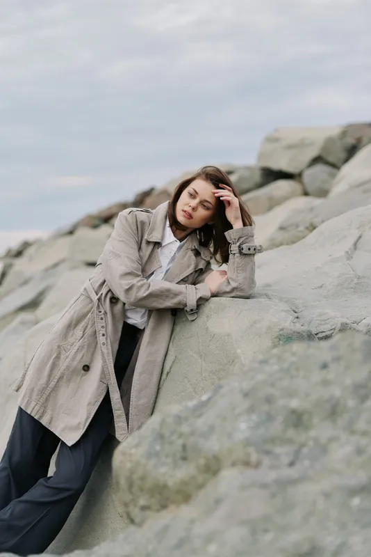 Young woman in textured coat leans on large rocks outdoors, looking contemplative. Moody sky and natural stone background create a calm and reflective atmosphere.